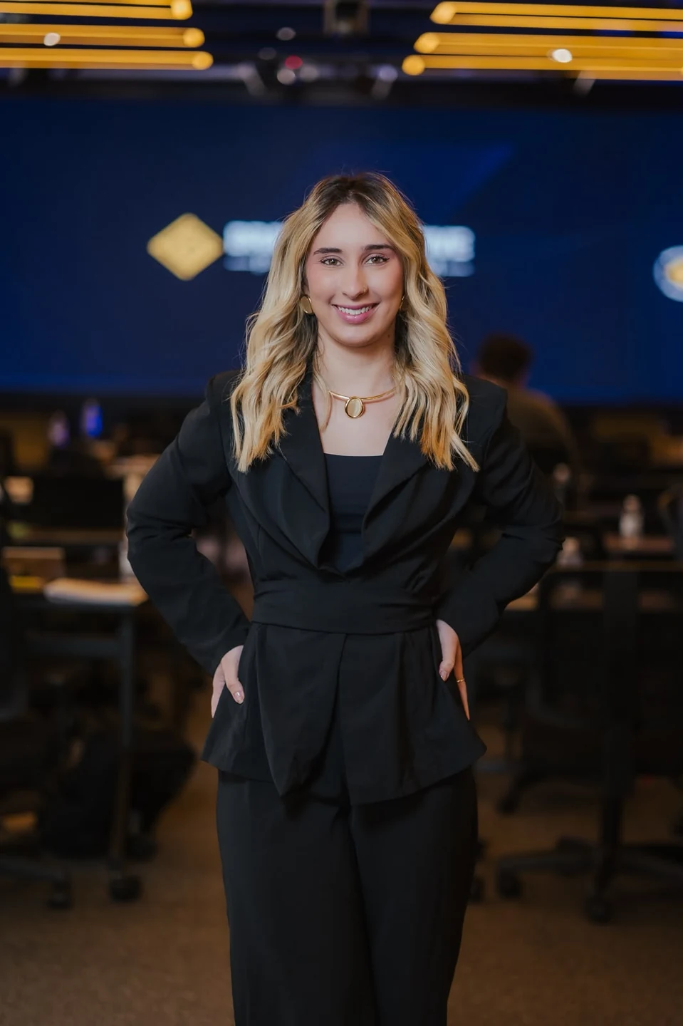 Camilly Parra, cabeleireira, sorrindo, com o cabelo loiro em ondas soltas e blazer preto